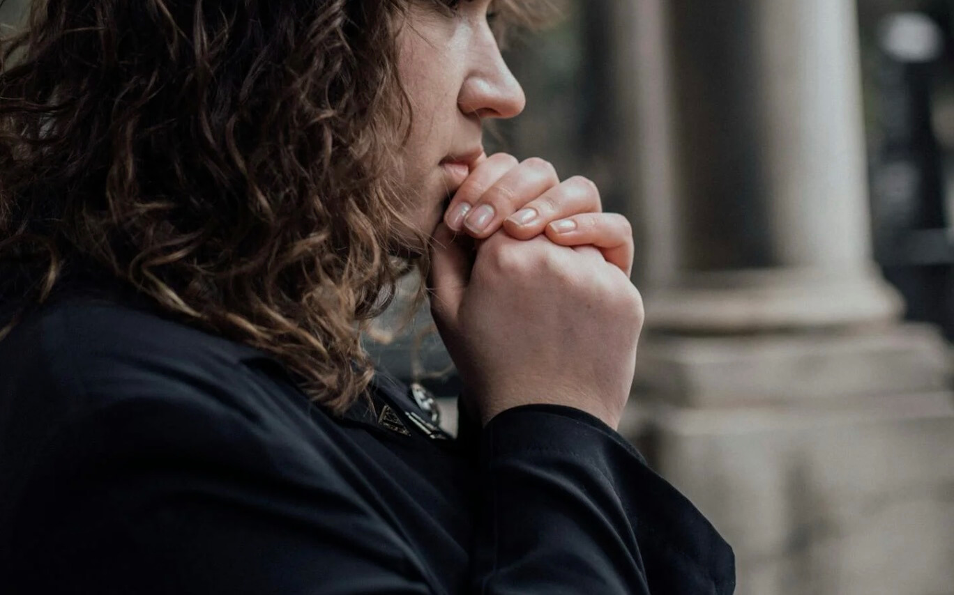 Close-up side profile of a grieving person with clasped hands, standing outdoors in a reflective moment.