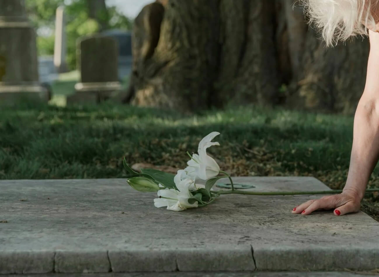 White flowers rest on a gravestone in a cemetery while a person’s hand touches the stone beside them.