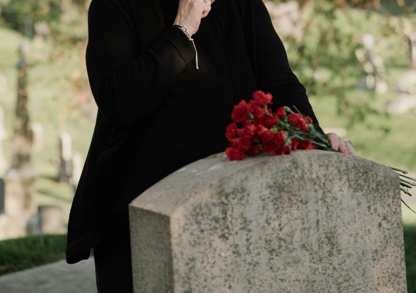 Person in black places red flowers on a gravestone in a cemetery during a quiet moment of remembrance.