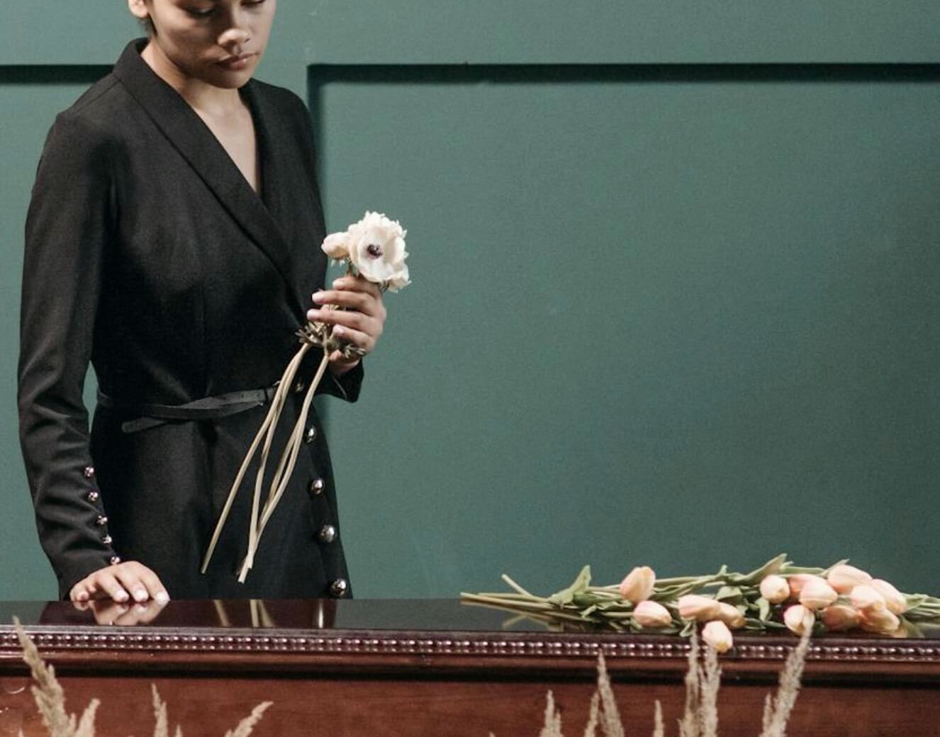Person in a black dress stands beside a polished wooden casket, holding pale flowers in a quiet funeral setting.